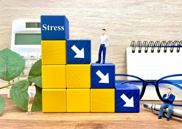 ストレス測定器の横に積まれた青と黄色のブロック、上部に「Stress」と書かれたブロックがあり、その上に小さなビジネスマンのフィギュアが立っている。机の上には眼鏡とメモ帳が置かれている。