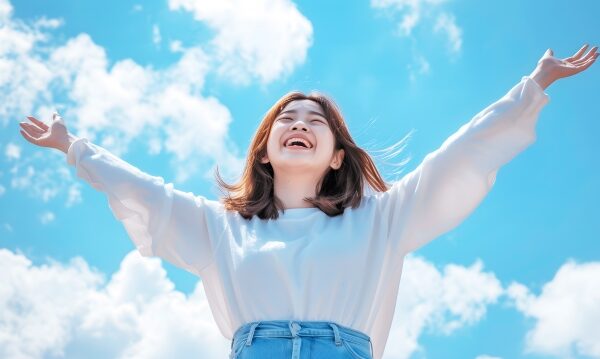 青空を背景に、両腕を広げて笑顔でいる女性。