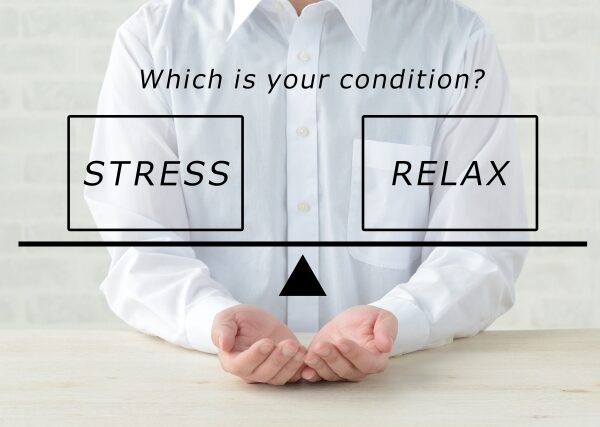 "A person wearing a white shirt with hands clasped on a desk, with two labels above reading 'STRESS' and 'RELAX' with a balance scale indicator in the middle."